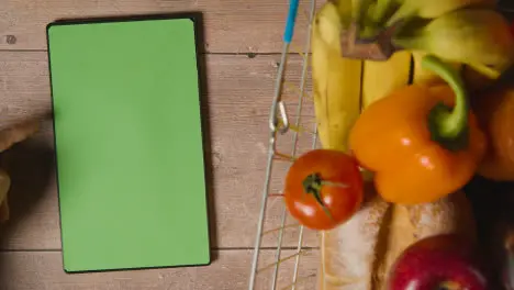 Overhead Studio Shot Of Person Using Green Screen Digital Tablet Next To Basic Food Items In Supermarket Wire Shopping Basket 1