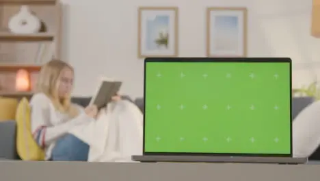 Woman Relaxing On Sofa Reading Book At Home With Green Screen Laptop In Foreground 