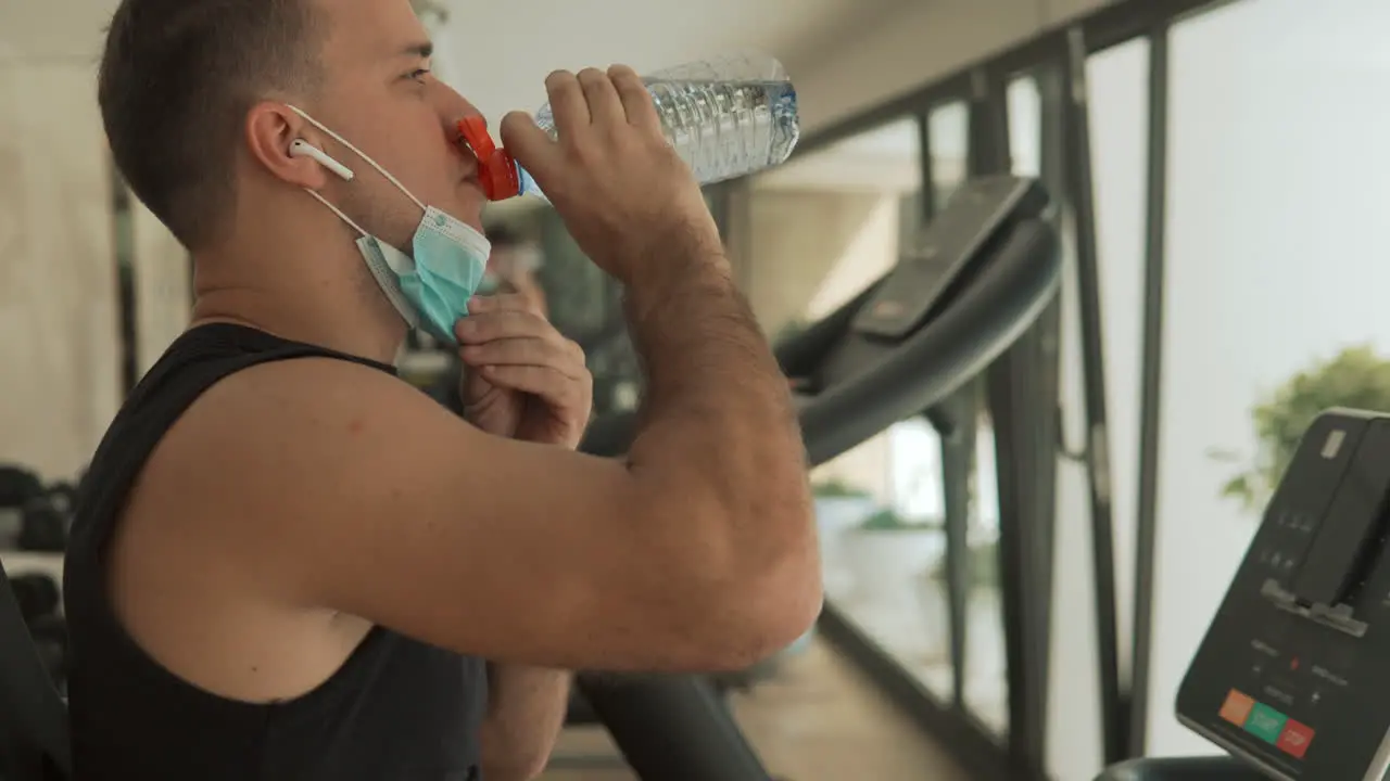 Young Strong Man With Face Mask Uses An Exercise Machine And Drinks Water In The Gym