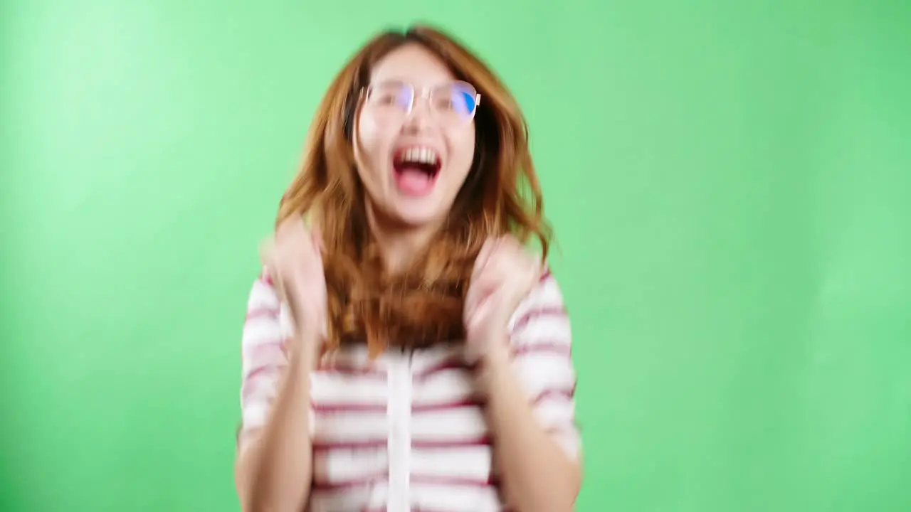 Cheerful young Asian woman with eyeglasses raising hands up shaking and having fist pump from happiness smiling woman face green screen background