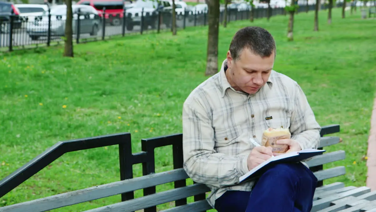 Man Eating Fast Food On A Park Bench Writes Something In A Notebook