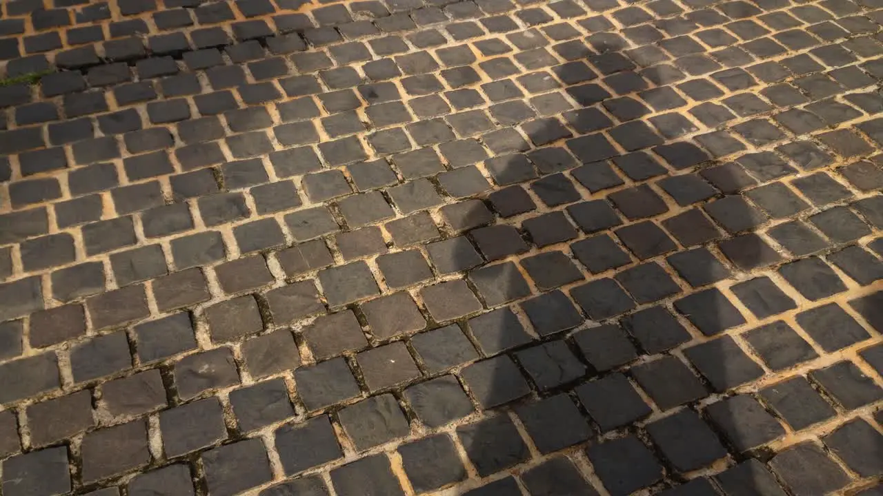 A man casting his own shadow while standing on a brick street pavement as other people pass by casting their own shadows as well