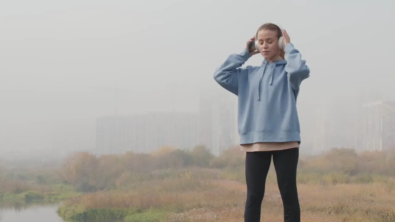 Young Serious Sportswoman In Blue Hoodie Putting On Headphones And Looking At The Horizon In The Woods On A Cloudy Day