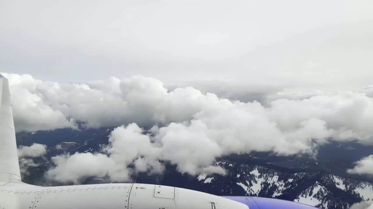 First person POV out of an airplane window as it flies over the mountains and through white puffy clouds