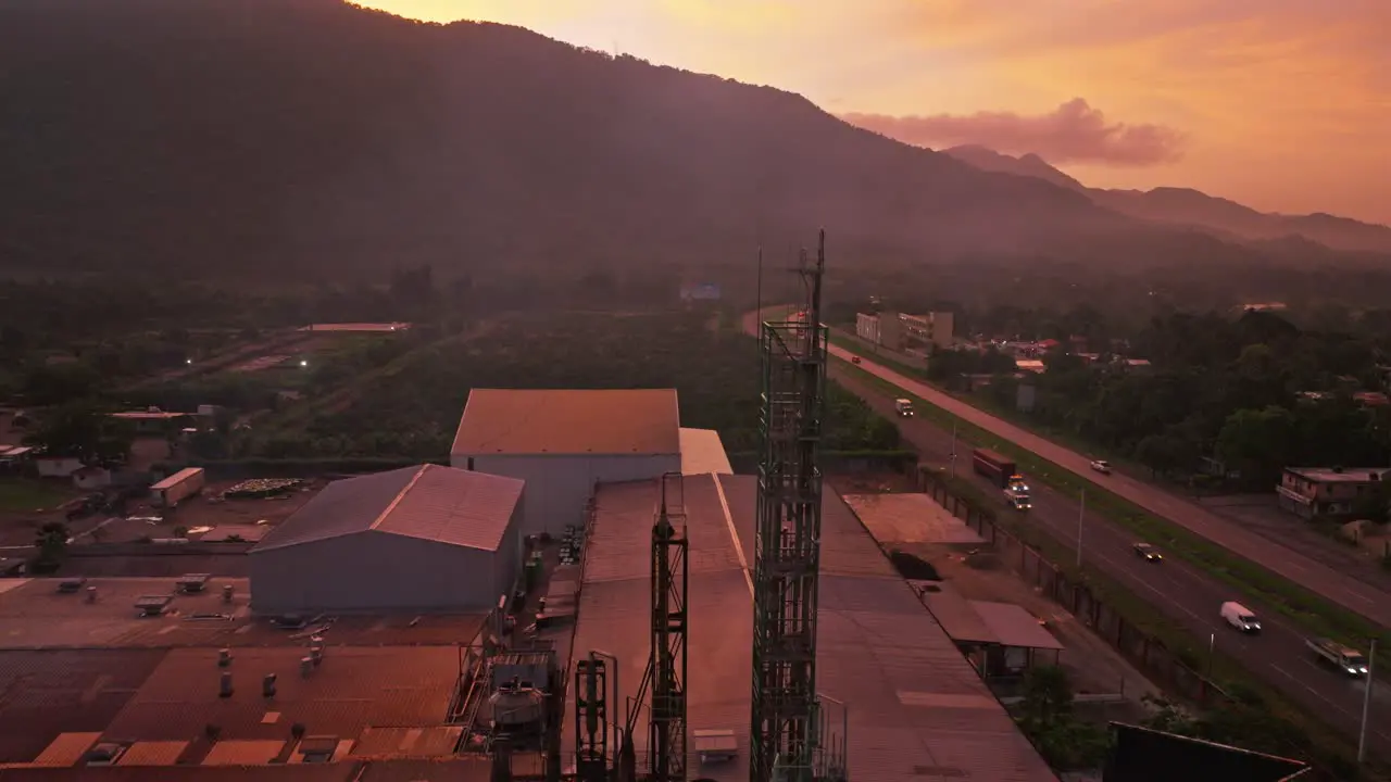 Ascending aerial shot of industrial factory and traffic on highway during golden sunrise behind mountains Villa Altagracia Dominican Republic