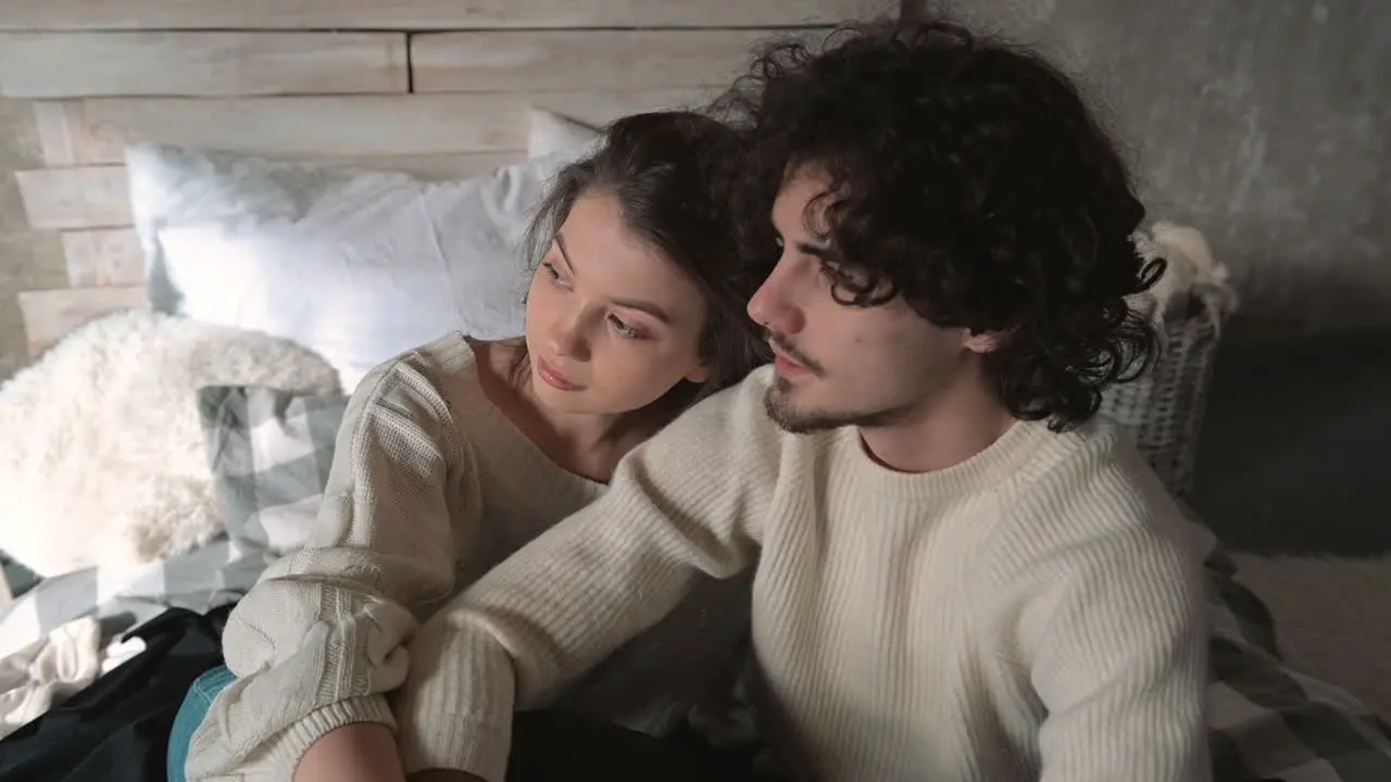 Young Romantic Couple Sitting In Bed Wearing Winter Clothes And Talking Hugging