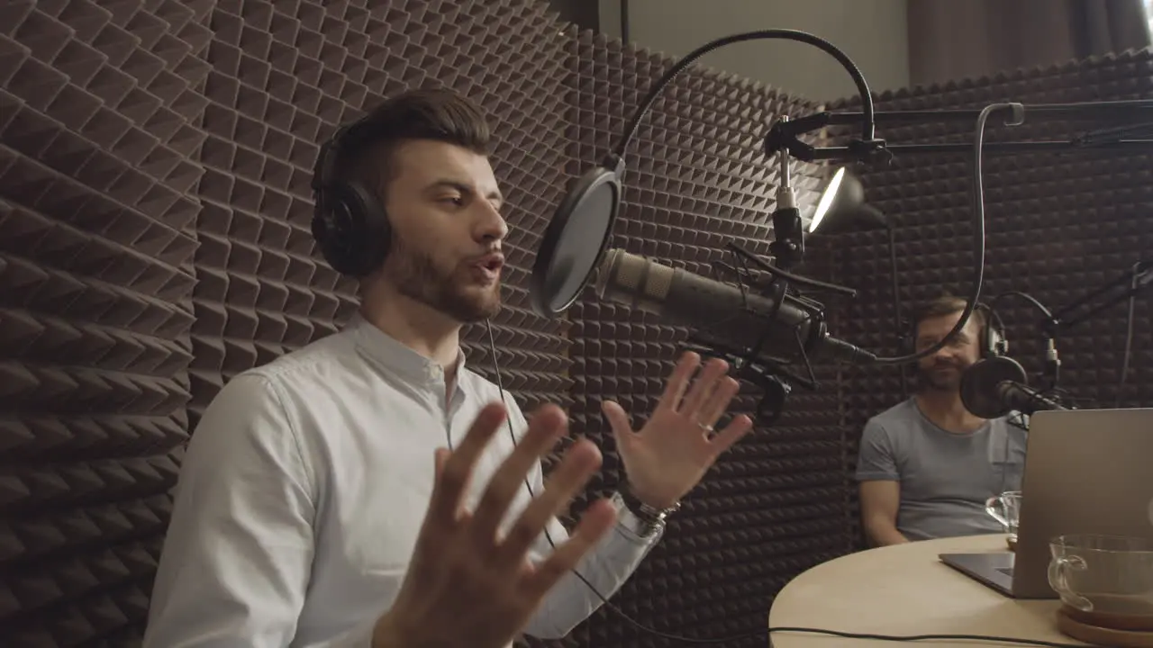 Bearded Young Radio Broadcaster Explains Something To A Person Sitting Next To Him In A Radio Recording Studio