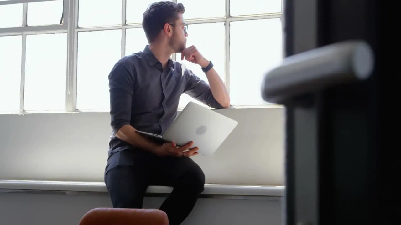 Front view of young caucasian businessman with laptop sitting near window in a modern office 4k