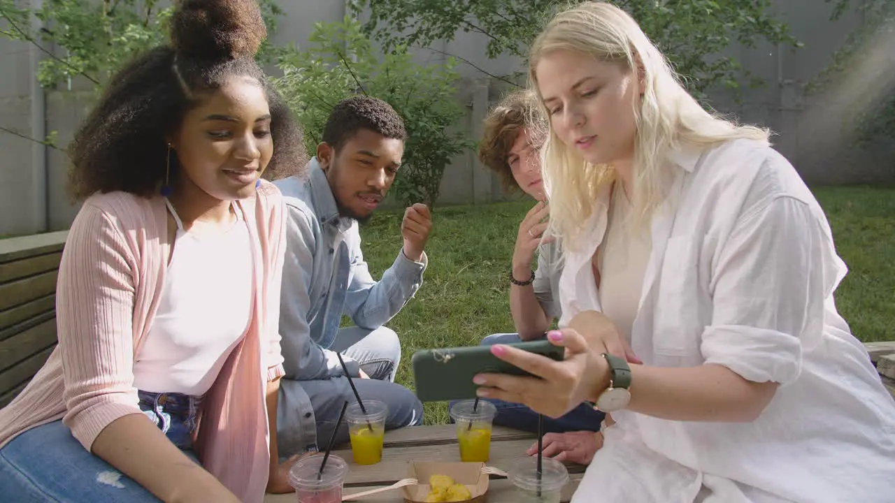 Young Blonde Woman Showing Something Interesting On Mobile Phone To Her Multiethnic Friends In A Park 1