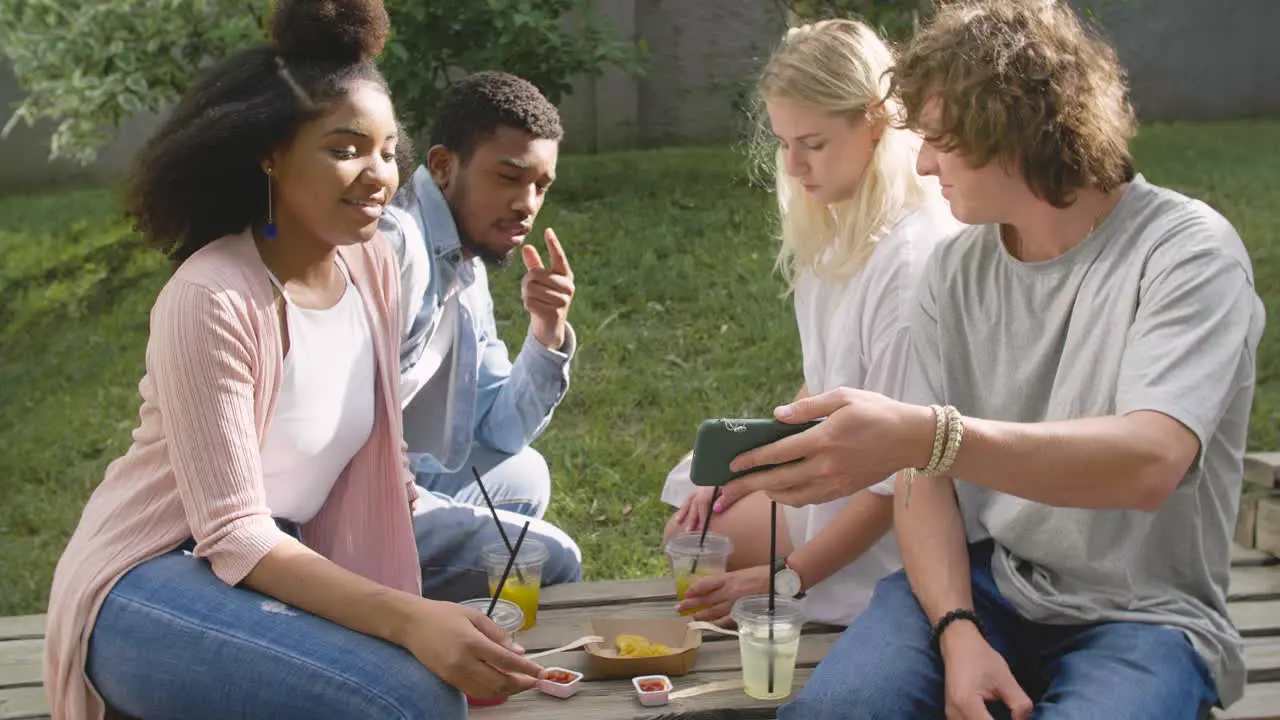 Young Man Showing Something Interesting On Mobile Phone To His Multiethnic Friends In A Park