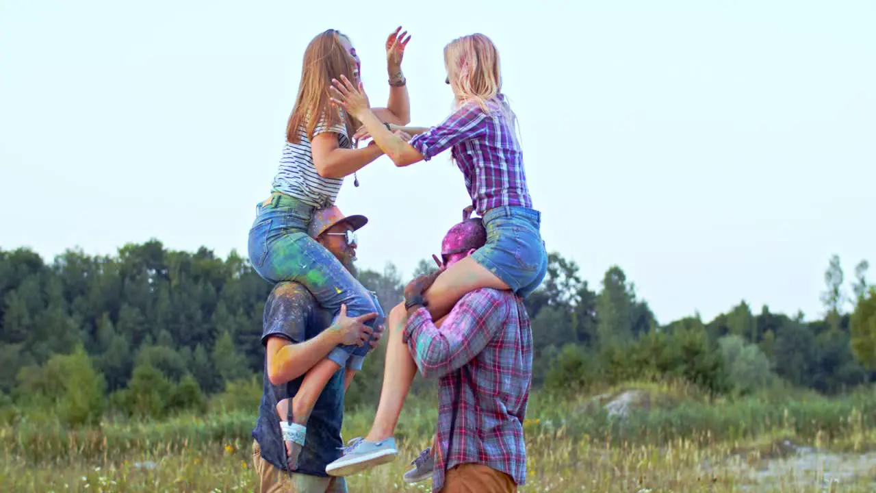 Two Blonde Happy Girls Sitting On The Shoulders Of Their Multiethnic Boyfriends And Having Fun At The Holi Holiday