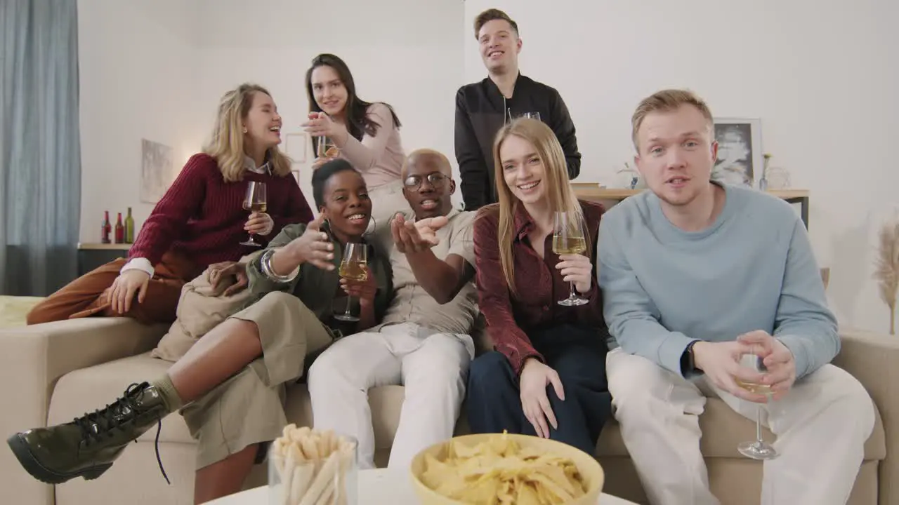 Group Of Friends Sitting On Sofa Drinking Wine And Waving At Camera At Home Party