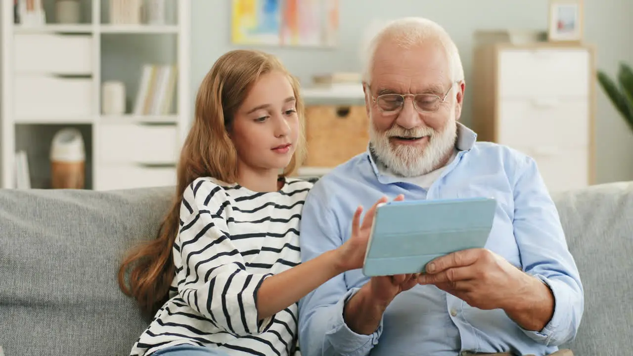 Portrait Of The Pretty Schoolgirl And His Grey Haired Grandfather In Glasses Who Showing Her Something On The Tablet Computer Screen Sitting On The Sofa At Home