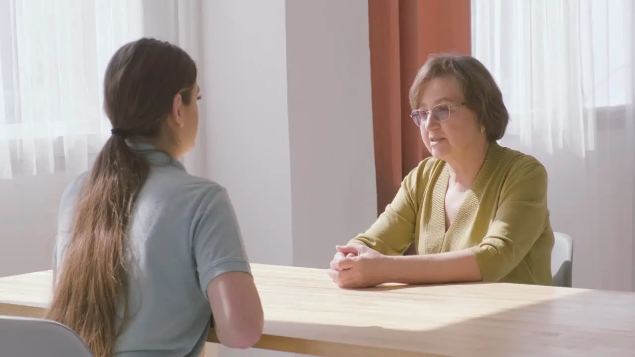 Senior Woman And Female Doctor Sitting On A Table Talking To Each Other 2