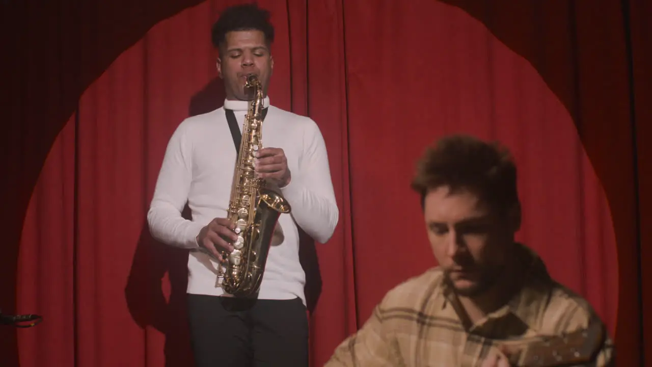 Latin Man Playing Sax And Man Sitting And Playing Guitar On Stage During A Rehearsal