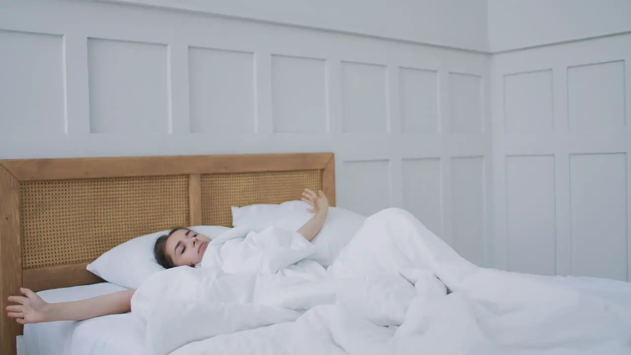 Young woman wakes up in bed in the morning smiling raises her hands