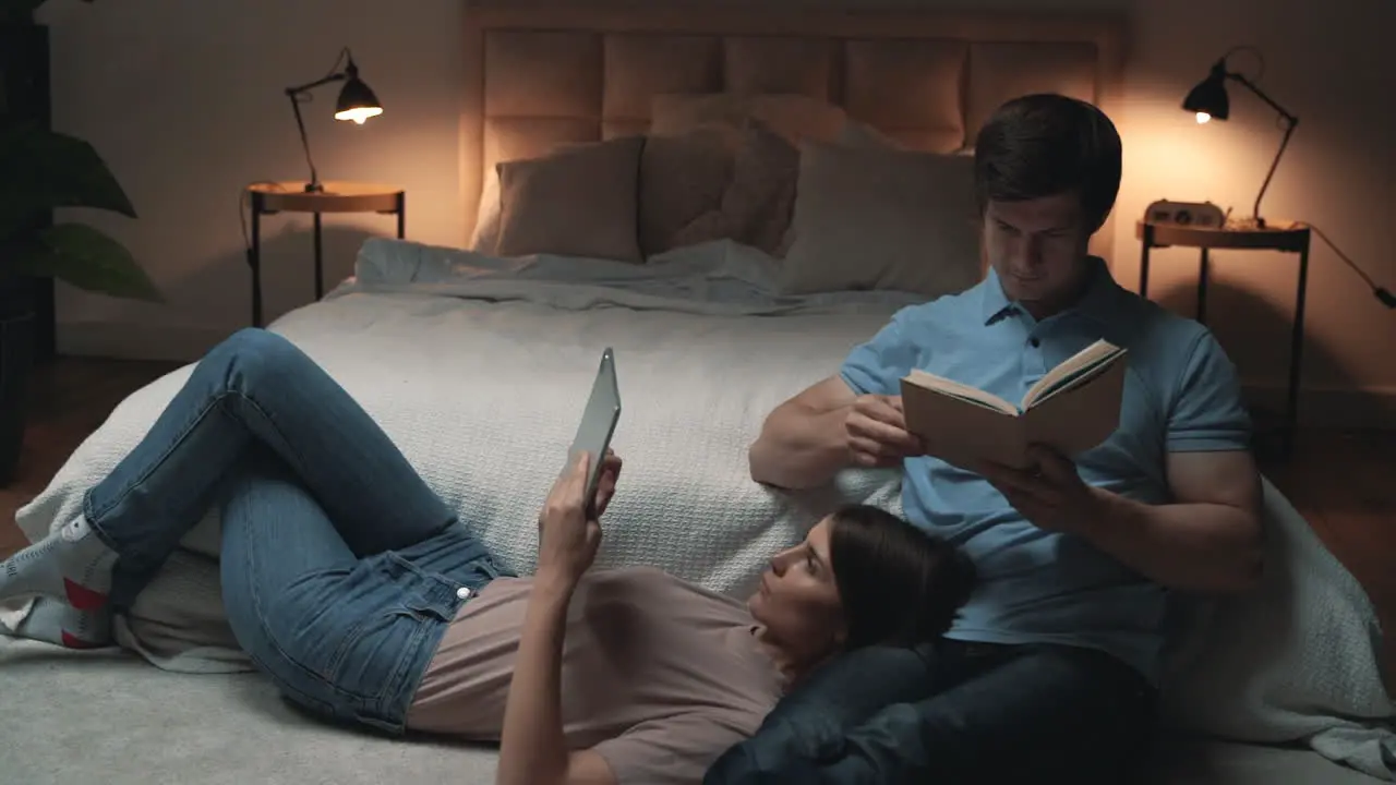 Young Couple Sitting In Bedroom Woman And Man Reading A Tablet And A Book And Relaxing Sharing Moments