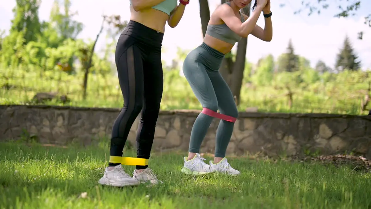 Two Fitness Women Doing Lateral Steps And Squats Exercises With Resistance Band In The Park