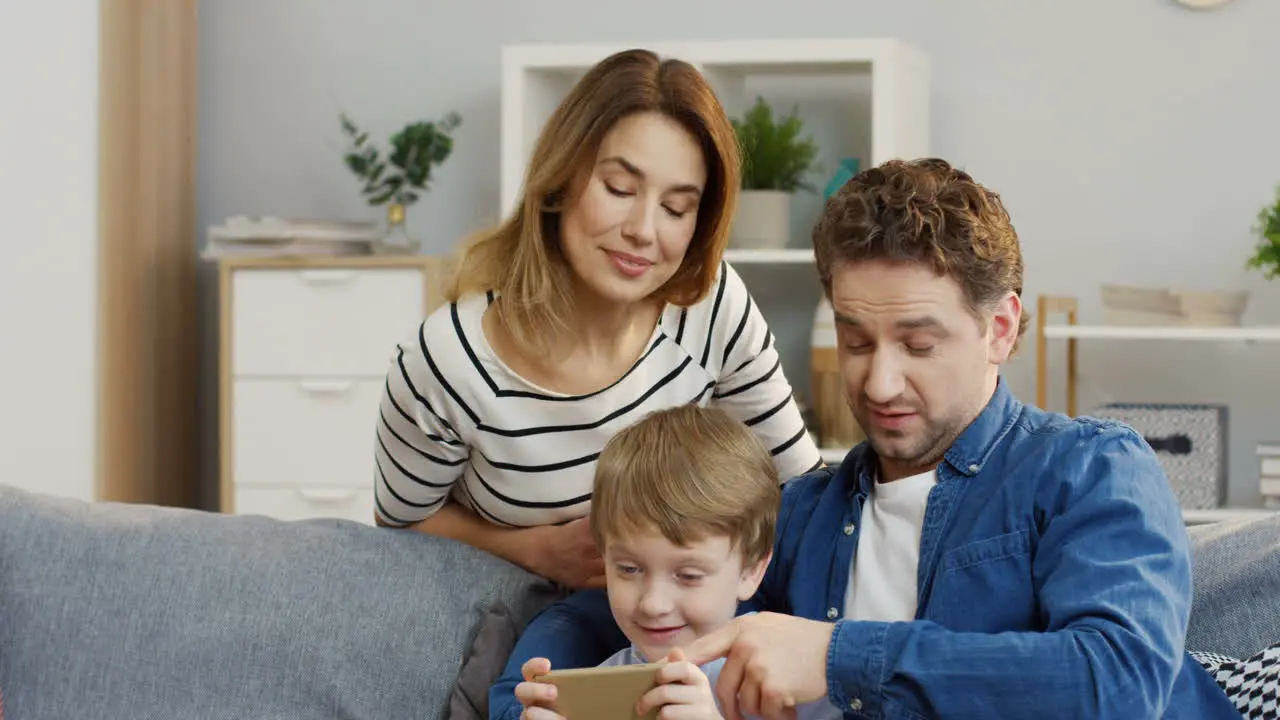 Little Cute Boy Sitting On The Sofa In The Living Room And Playing Games On The Smarthone His Parents Watching Him