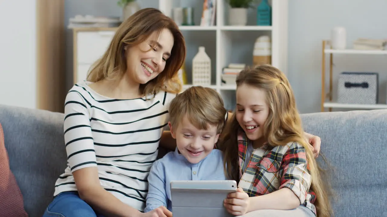 Portrait Of The Pretty Mother Sitting On The Gray Couch And Hugging Her Cute Small Son And Teen Daughter While They Watching Something On The Tablet Computer In The Living Room