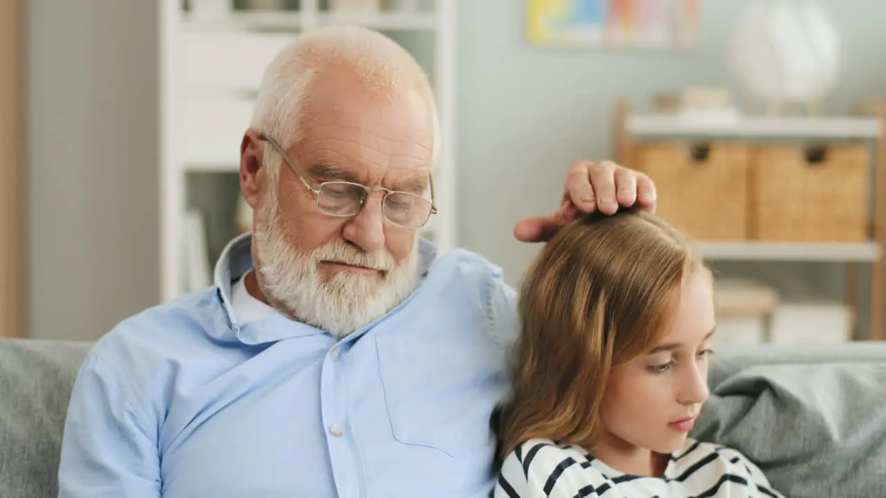 Close Up Of The Old Grandpa Reading A Book And Caressing His Granddaughter On The Head While She Sitting Beside With A Smartphone