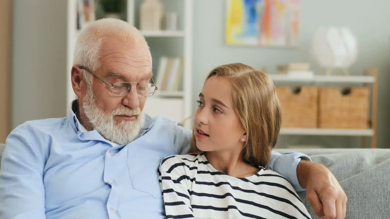 Portrait Of The Smiled Grandpa In Glasses And Blonde Pretty Grandaughter Sitting Close Together On The Sofa In The Cozy Room