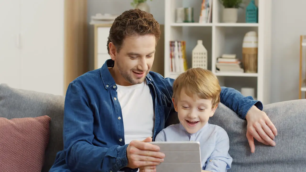 Handsome Man Sitting On The Gray Sofa And Hugging His Cute Little Son When They Watching Something On The Tablet Computer And Talking In The Living Room