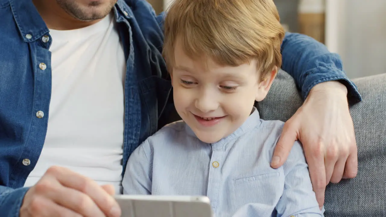 Close Up Of The Attractive Father Sitting On The Couch And Hugging His Small Son While They Watching Something On The Tablet Device