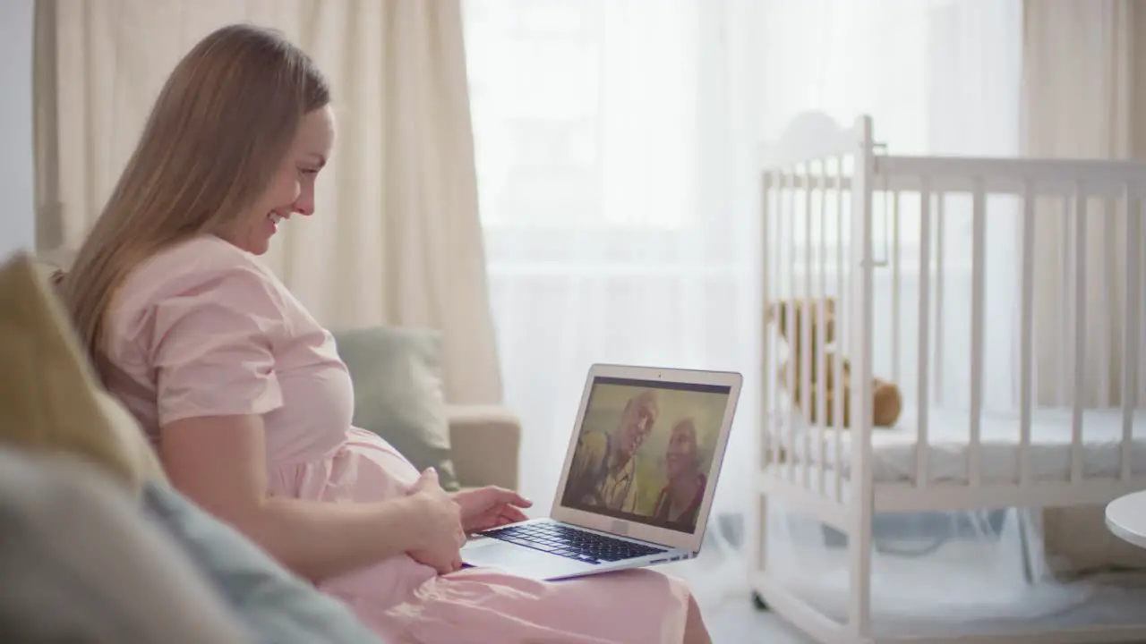 Pregnant Woman Sitting On Sofa And Using A Laptop Making A Video Call With His Parents