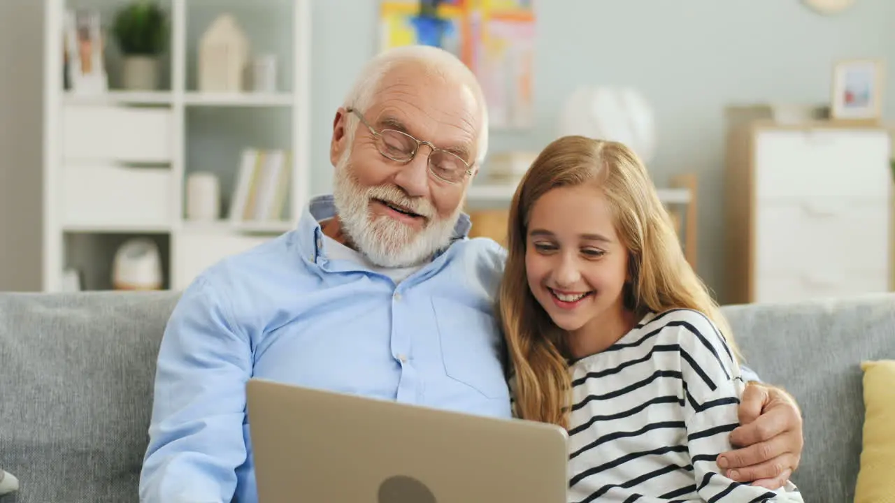 Portrait Shot Of The Pretty Teenage Girl Laughing Together With Her Grandfather While They Looking At The Screen Of Laptop Computer In The Nice Living Room