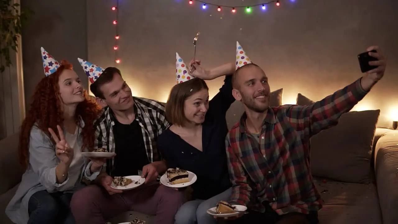 Cheerful Young Friends Or Two Couples Having Small Cozy Birthday Celebration Sitting Together In Festive Cones On The Couch And Taking Selfie Using A Mobile Phone 1