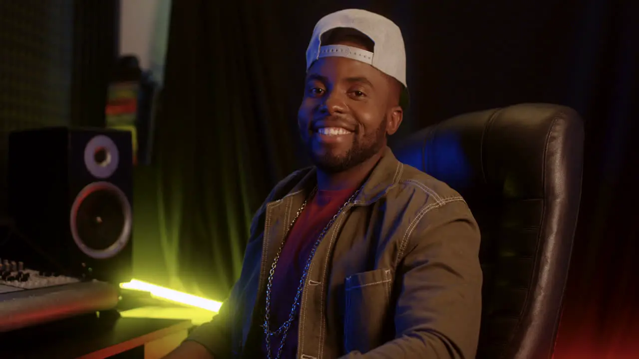 Handsome Young Man Sound Producer In A Hat Sitting In The Music Studio At The Mixer And Working Then Turning Head To The Camera And Smiling Cheerfully