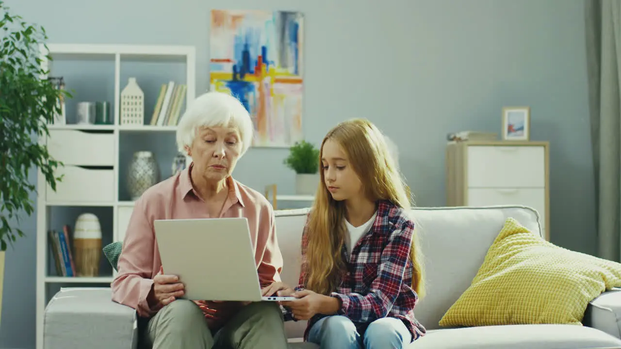 Old Good Looking Woman Sitting On The Couch In The Cozy Livingroom And Telling Something To Her Pretty Teen Granddaughter While Demonstrating Something On The Laptop Screen