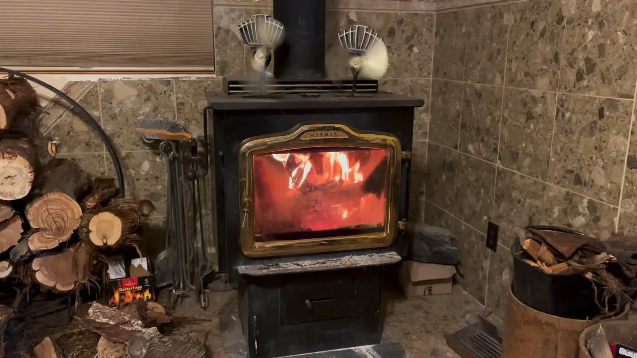 A wood burning stove with firewood stacked next to it burns strong with two kinetic fans sitting on top of the stove