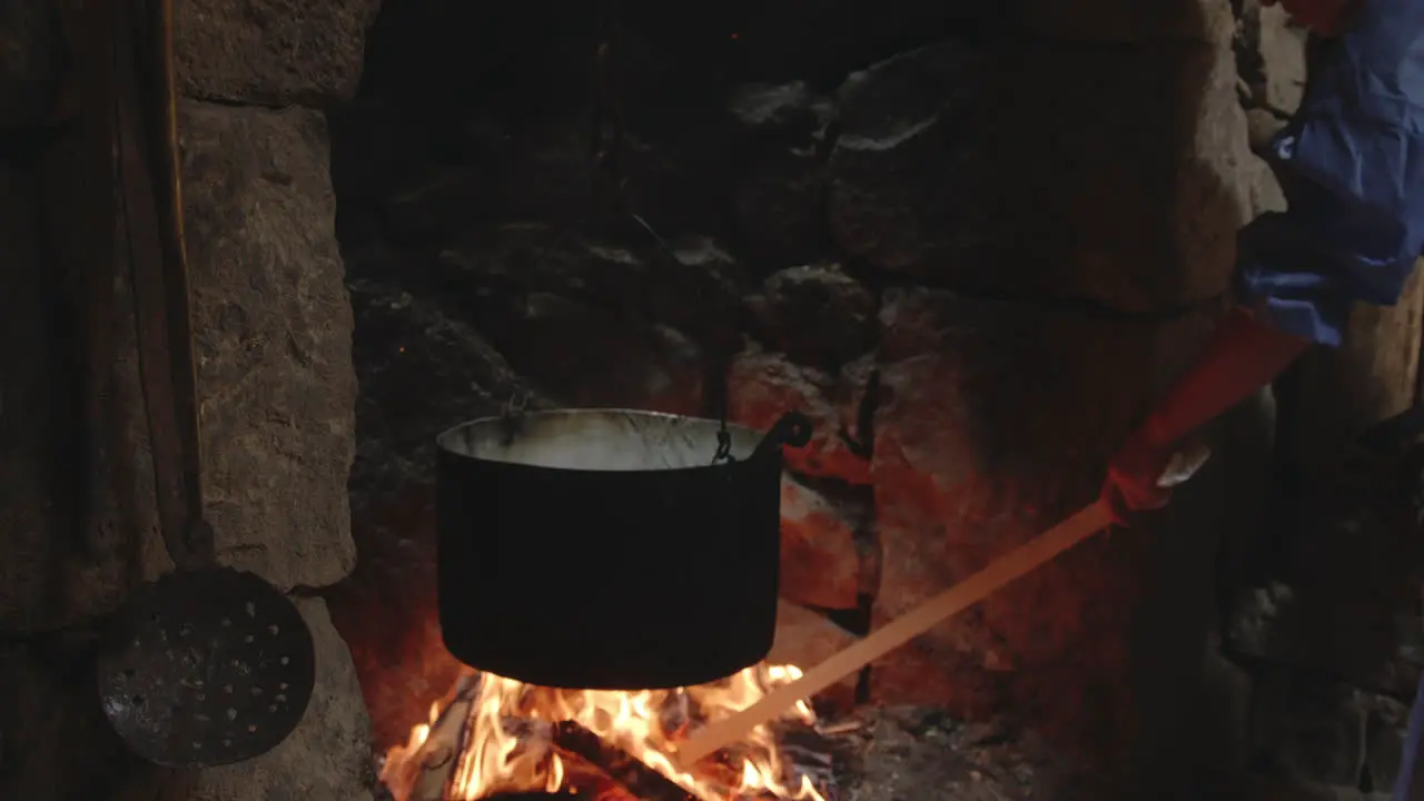 Woman raking buring logs in flames below cauldron in stone fireplace