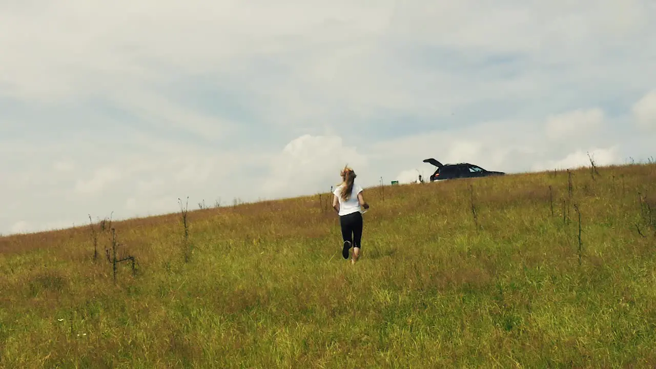 Long haired blonde girl running up a hill on grass in slow motion with clouds in the background drone follow cam