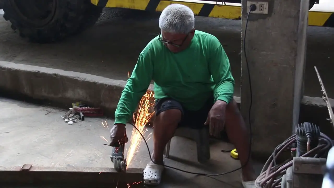 wideshot of an old man with white hair cutting a metal bar using an angle grinder with no protective guards like face shield gloves shoes and working suit