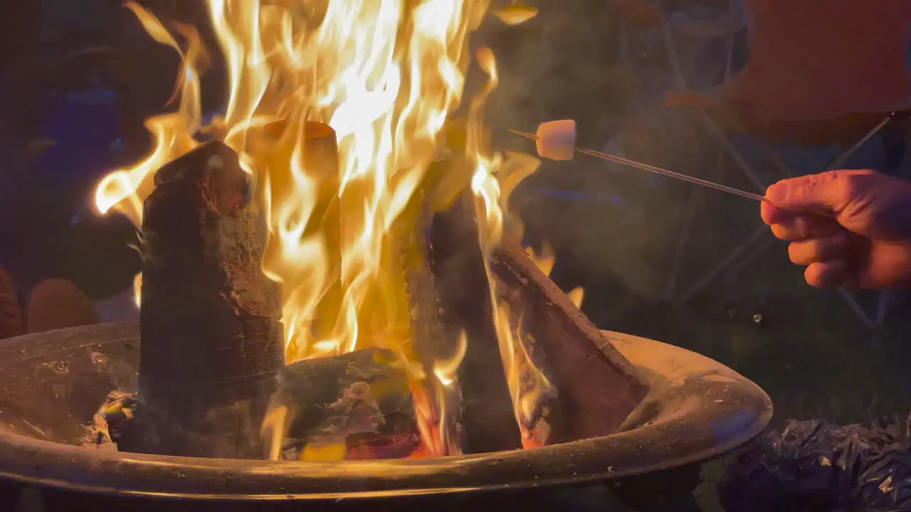 Static shot of a camp fire burning with people sitting round cooking marshmellows