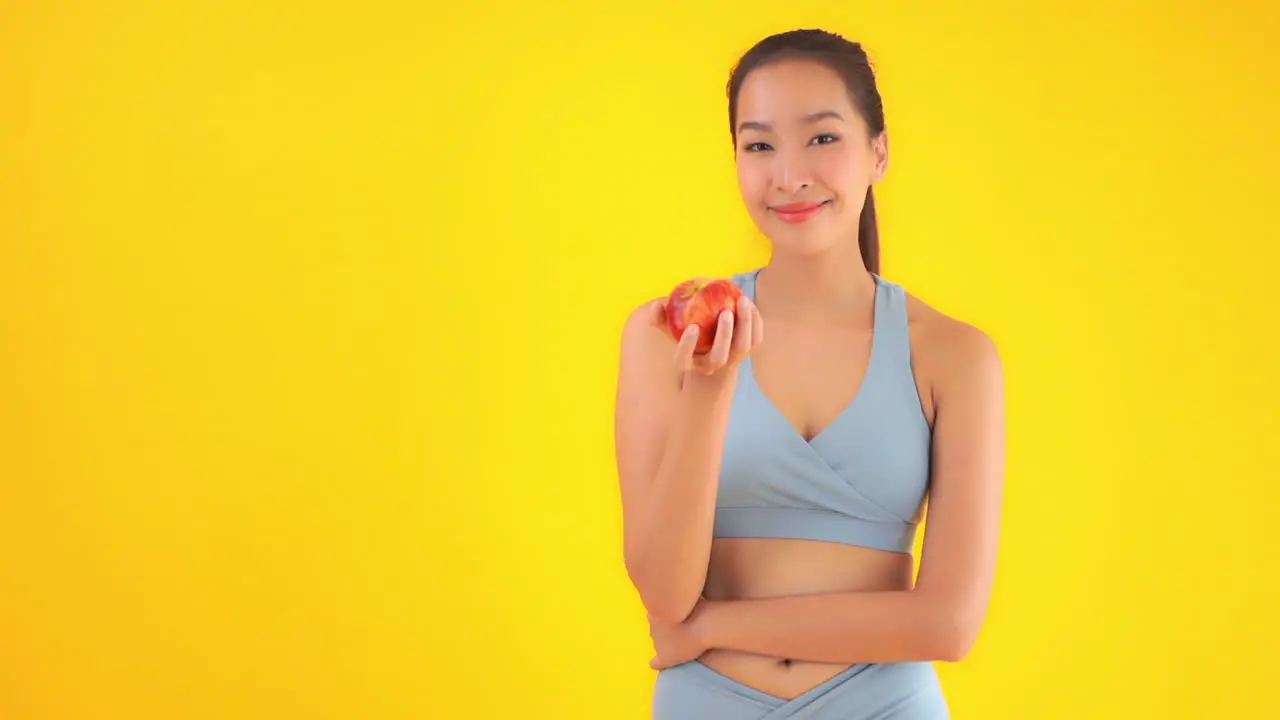 Asian Female in Gym Sportswear With Fresh Red Apple Slow Motion Healthy Food and Lifestyle Concept Static Shot Isolated on Yellow Background