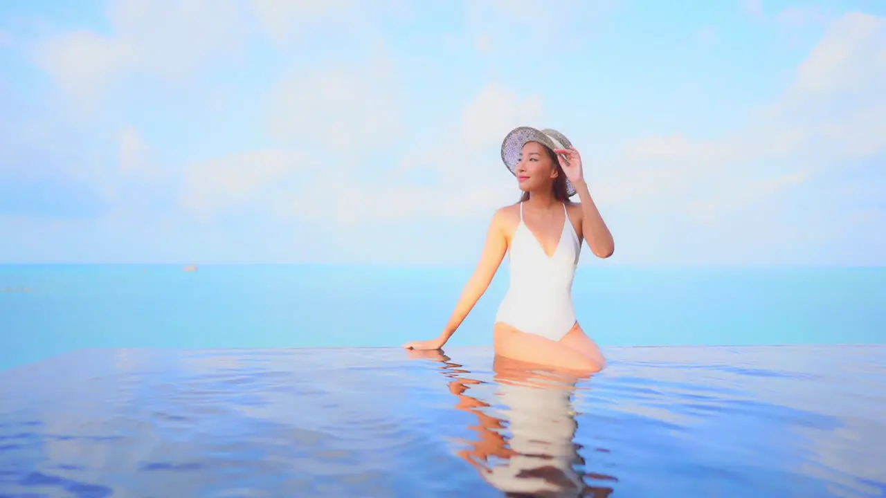 A pretty young woman in a one-piece bathing suit sits on the edge of an infinity pool with an ocean background