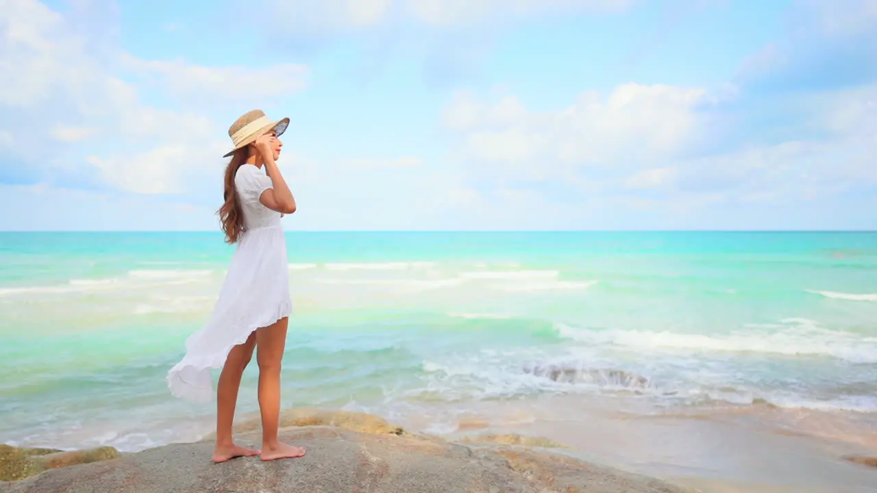 A young woman in a sundress holds on to her sun hat as she overlooks the incoming waves