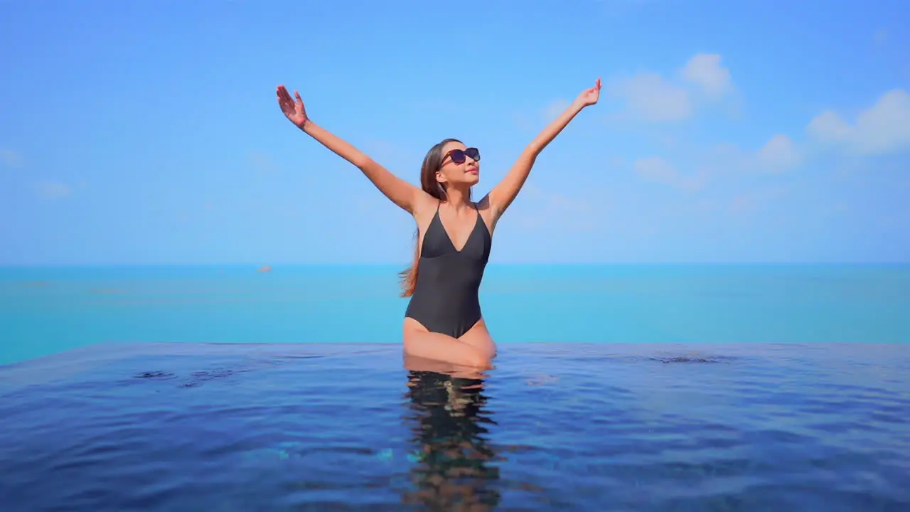 With a small boat on in the ocean background a pretty young woman is sitting on the invisible edge of a swimming pool raises her arms in welcome to the glorious weather