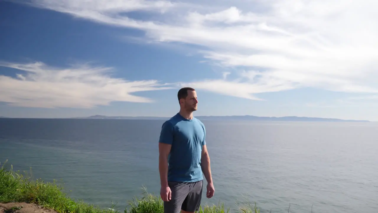 A fit strong man standing proud on the edge of an ocean cliff nature hiking trail with an epic beach view on a sunny day in Santa Barbara California SLOW MOTION