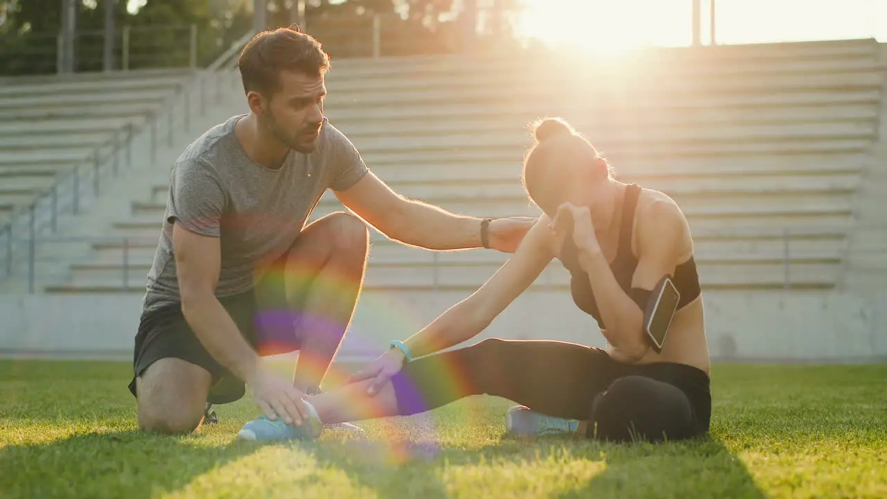 Young Jogger Woman Sitting On Green Grass And Having Pain In Her Leg As Injured During Workout In The Stadium On A Sunny Day 1