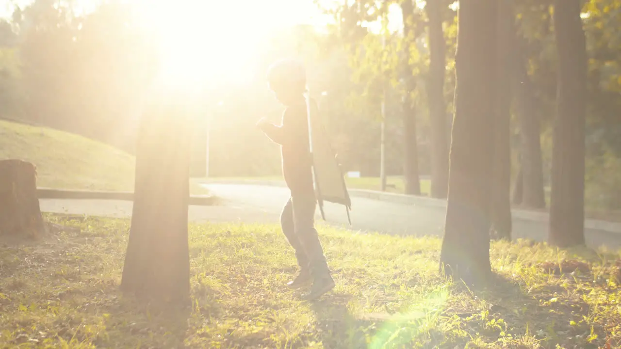 Little Boy In Helmet And Red Sweater With Cardboard Airplane Wings Running Around The Trees In The Park On A Sunny Day And Playing As A Pilot