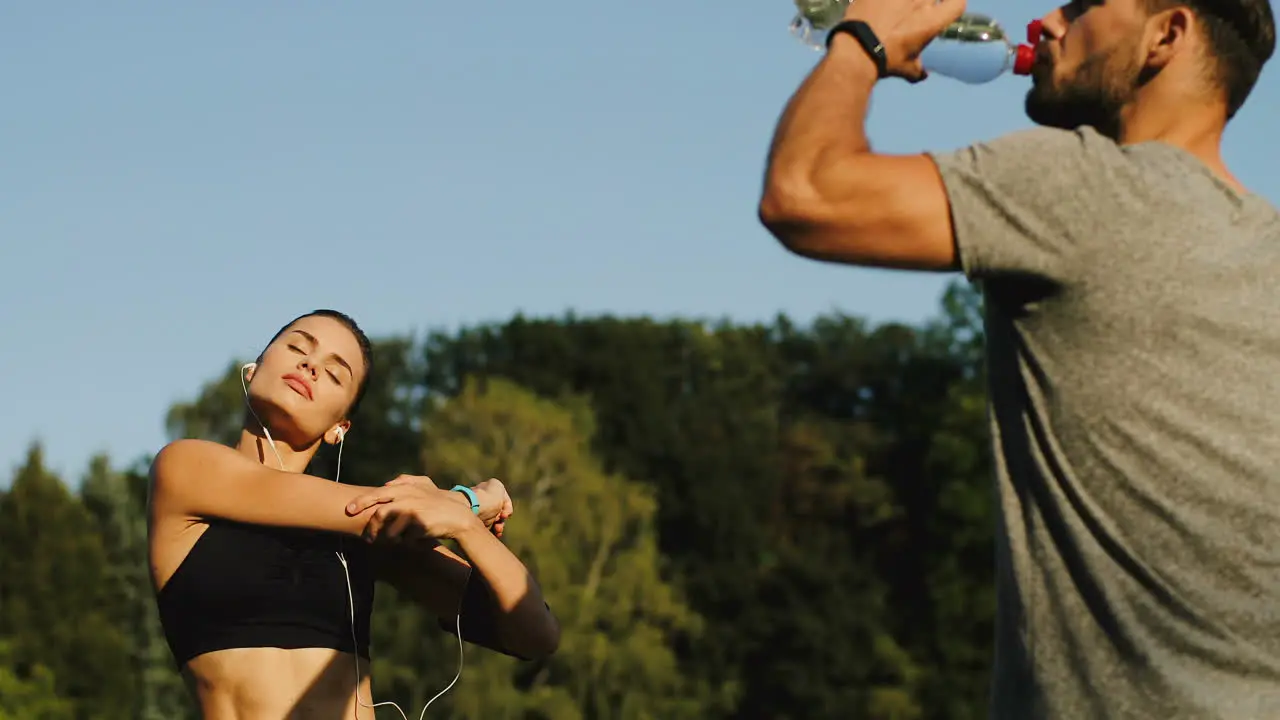 Rear View Of Jogger Man Drinking Water And Young Woman In Headphones Exercising And Warming Up Hands And Shoulders On A Sunny Summer Day