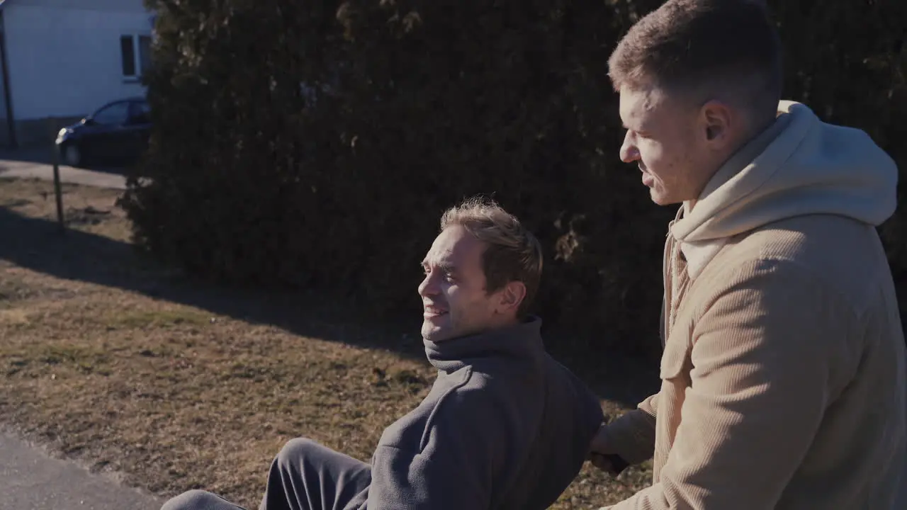 Young Man Walking While Pushing His Disabled Friend In Wheelchair And Talking Together