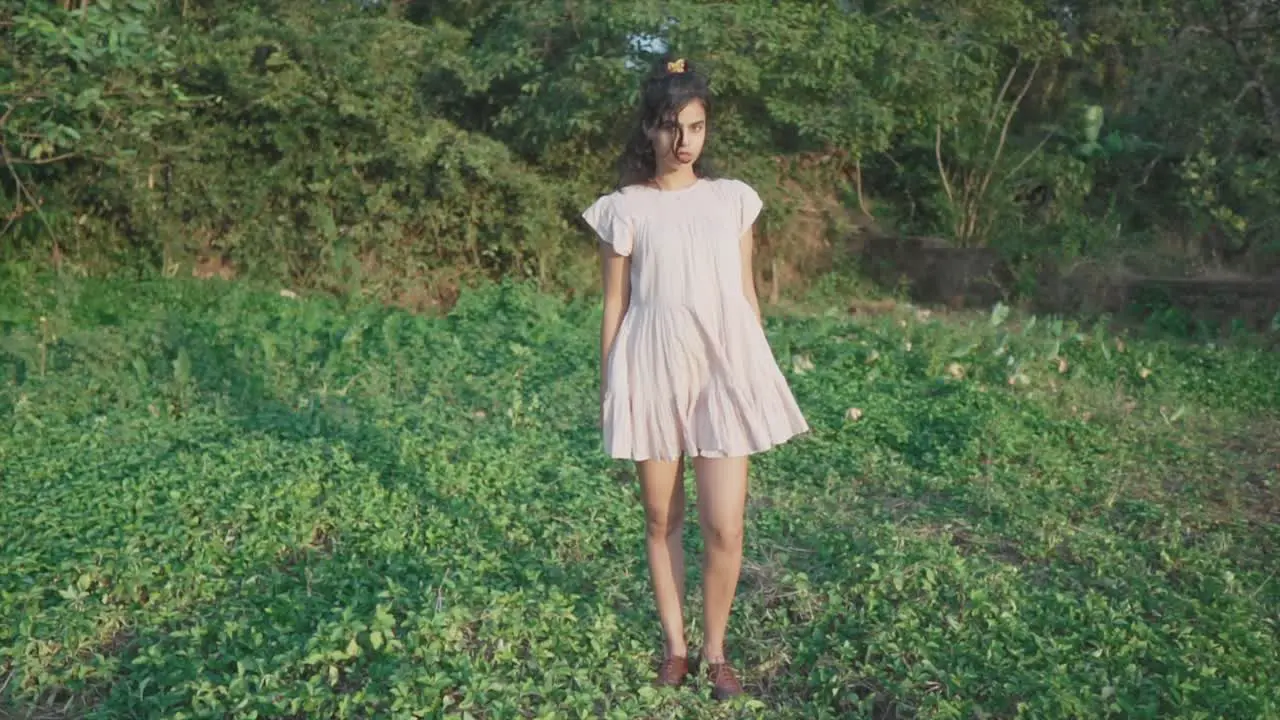 Slow motion static shot of a young pretty indian woman dressed in a pink dress standing in the garden in the grass between trees in the background looking at the camera