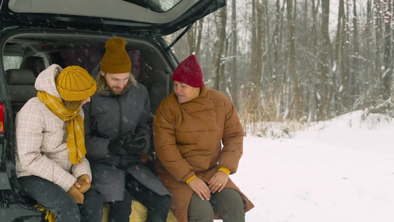 Bearded Man Sitting With Two Friends In Car Boot And Looking At Mobile Phone On A Winter Day