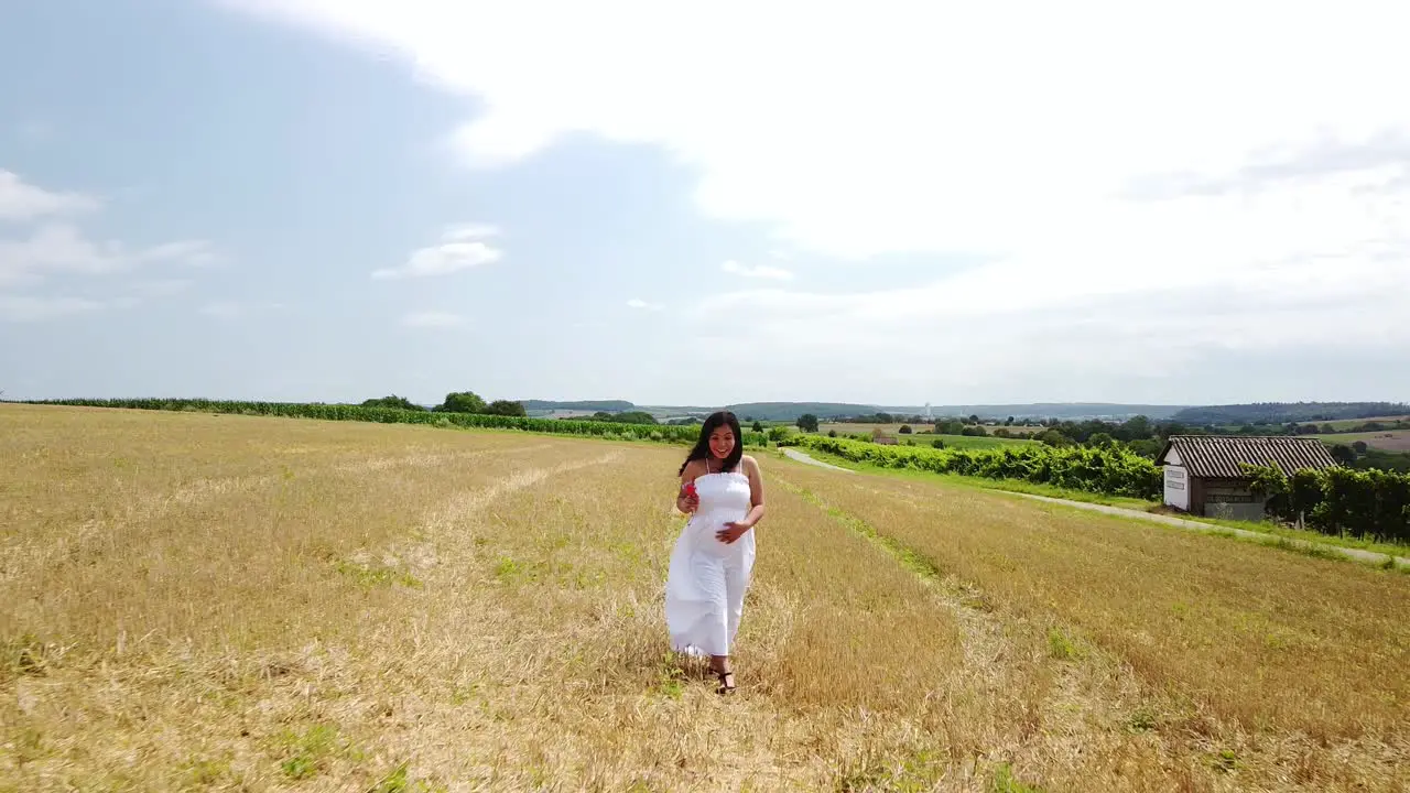 Young pregnant Woman in a White Summer Dress walking down a field holding her belly and flowers for a Maternity Photo Shoot